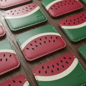 Könnte beinhalten: Eine Sammlung von Kartenhaltern im Wassermelonen-Design. Jeder Kartenhalter hat ein rotes, grünes und weißes Design mit schwarzen Samen-Details. Die Kartenhalter sind rechteckig und aus einem lederähnlichen Material gefertigt. Der Text "mugi mugi goods" ist sichtbar.