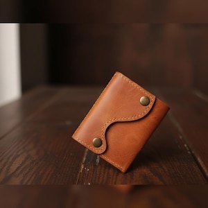 Op de afbeelding: Een kleine, bruine leren portemonnee met een gebogen flap die is vastgezet met twee messingkleurige drukkers. De portemonnee is rechthoekig met zichtbare stiksels en staat op een donkere houten achtergrond.