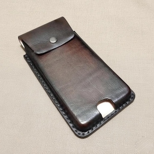 May include: A dark brown leather phone case with a flap closure and a snap button. The case has a rectangular shape with a stitched border and a cutout at the bottom. The leather has a slightly distressed appearance.