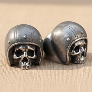 May include: Two silver-toned metal rings, each featuring a detailed human skull wearing a vintage-style motorcycle helmet. The helmets have a textured, matte finish with visible rivets along the top and sides, while the skulls show deep eye sockets.