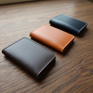 May include: Three leather cardholders in a row, each in a different colour. The first is dark brown, the second is tan, and the third is navy blue. Each cardholder has a stitched border. The cardholders are on a wooden surface.