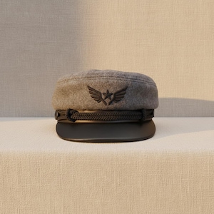 May include: A gray military-style cap with a black brim and a decorative rope detail. The cap features a black emblem of a star within wings. This hat is a stylish accessory.