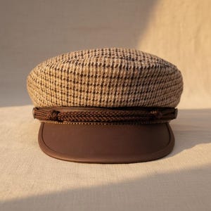 Peut inclure: Casquette de marin marron et beige à motif pied-de-poule, avec une visière en cuir marron foncé. Le chapeau est orné d'un détail de corde tressée. Un accessoire de mode classique et élégant.