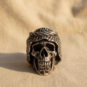 Puede incluir: Anillo de calavera plateado con un diseño detallado. La calavera lleva un casco con un patrón de cota de malla y una banda trenzada. El anillo tiene una estética gótica detallada.