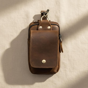 Könnte beinhalten: Eine braune Gürteltasche aus Leder mit einem rechteckigen Hauptfach mit Reißverschluss und einer Fronttasche mit Klappe, die durch einen einzelnen Metalldruckknopf gesichert ist. Die Tasche verfügt über einen D-Ring aus Metall zur Befestigung.