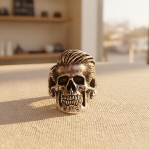May include: A silver-colored skull ring with detailed facial features and a sculpted hairstyle. The ring is displayed on a neutral-toned surface, showcasing the intricate design and craftsmanship of the jewelry.
