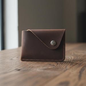 May include: A brown leather wallet with a snap closure. The wallet is rectangular with rounded corners and a stitched border. The snap is silver-colored. The wallet is sitting on a wooden surface.