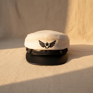 May include: Cream-colored cap with a black brim and a black rope detail. The cap features a black embroidered emblem of a star with wings. The hat is sitting on a beige surface.