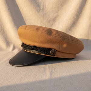 Puede incluir: Una gorra de marinero color beige con visera negra y detalle de cuerda negra. El sombrero presenta un pequeño botón redondo con un diseño de ancla. El sombrero está hecho de tela texturizada.