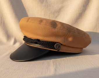 May include: A tan sailor hat with a black brim and a black rope detail. The hat features a small, round button with an anchor design. The hat is made of a textured fabric.