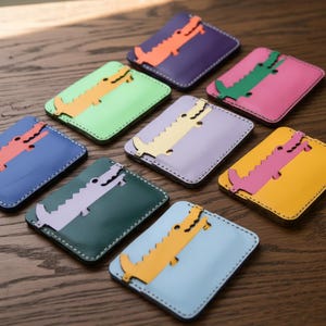 Puede incluir: Colección de tarjeteros de cuero de colores, cada uno con un diseño de caimán estilizado. Los tarjeteros vienen en varios colores, incluyendo azul, verde, morado, rosa y amarillo. El caimán es de un color contrastante.