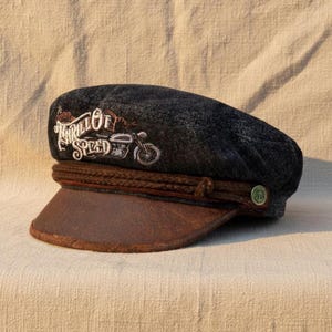 Puede incluir: Gorra de mezclilla oscura con visera de cuero marrón y banda trenzada. La gorra presenta texto bordado y un diseño de motocicleta. Un pequeño botón verde con un emblema náutico está en el lateral.