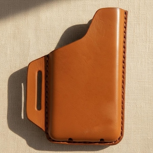 Könnte beinhalten: Eine hellbraune Leder-Handyhülle mit Gürtelschlaufe. Die Hülle hat eine rechteckige Form mit abgerundeten Ecken und ist an den Kanten genäht. Das Leder hat eine glatte, polierte Oberfläche.