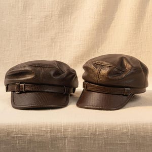 May include: Two brown leather newsboy caps with a textured finish. Each cap has a short brim and a decorative strap with a buckle. The caps are displayed on a neutral-coloured surface.