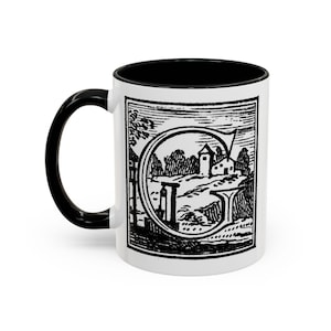 Può includere: Tazza in ceramica bianca con manico e interno neri. La tazza presenta un'illustrazione in bianco e nero di un paesaggio con un edificio, alberi e la lettera "G".
