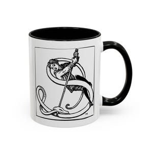 Può includere: Tazza in ceramica bianca con interno e manico neri. La tazza presenta un'illustrazione in bianco e nero di una persona che tiene un palo, intrecciata con una lettera "S" stilizzata e un serpente.