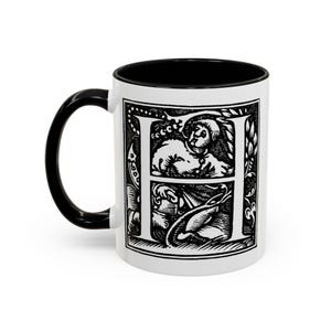 Puede incluir: Taza de cerámica blanca con asa e interior negros. La taza presenta una detallada ilustración en blanco y negro de la letra "H" con un diseño de estilo medieval, que incluye figuras y elementos florales. Un artículo decorativo para el hogar o la oficina.