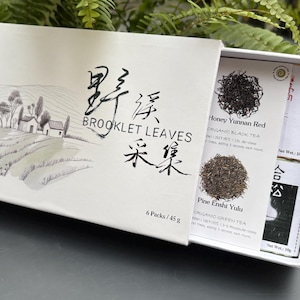 Puede incluir: Una caja de té blanca con el texto "BROOKLET LEAVES" y caracteres chinos. La caja contiene paquetes de té individuales etiquetados como "Honey Yunnan Red" y "Pine Enshi Yulu". La caja tiene una ilustración de paisaje.
