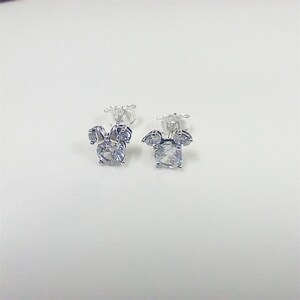 May include: A pair of silver-toned stud earrings. Each earring features a large, round, clear gemstone at the center, surrounded by smaller, clear gemstones arranged in a bow shape. The earrings are set against a white background.