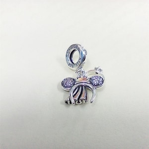 May include: A silver charm with a Mickey Mouse-inspired design. The charm features a pair of round, gem-studded ears and a bow. The charm is attached to a ring with the text "925" on it.