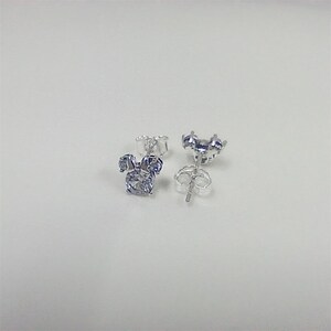 May include: A pair of silver-colored stud earrings. Each earring features a three-part design, with a large, round, light blue stone at the center and two smaller stones on top, resembling a cartoon character's head. The earrings are on a white surface.