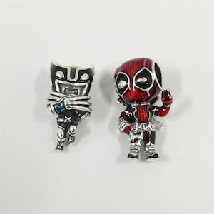 Puede incluir: Dos dijes de metal con figuras de personajes estilizados. El dije de la izquierda es una figura negra y plateada con máscara y manos en forma de garra. El de la derecha es una figura roja y negra con máscara texturizada y mano levantada.
