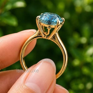Puede incluir: Un anillo de oro con una gran piedra preciosa redonda y facetada de color azul claro en un engaste de garras alto. El lateral de la galería de oro muestra una letra J prominente. El aro es liso y pulido, con un acabado metálico clásico.
