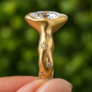 May include: A gold ring with a textured, organic band featuring four small star-shaped engravings, each set with a tiny clear gemstone. The ring is topped with a large, faceted, clear oval stone. The text ISHVALUXE is visible at the base.