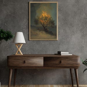 May include: A framed art print depicts a tree engulfed in flames, with a dark, moody sky. The artwork is set against a gray wall, above a wooden console table with a lamp and books.