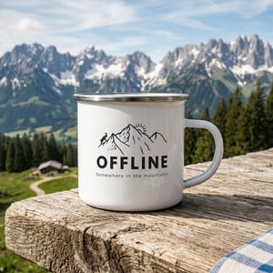 Könnte beinhalten: Ein weißer Emaillebecher mit silbernem Rand und Henkel, der eine schwarze Grafik von Berggipfeln, einer kleinen Ziege und einer Sonne zeigt, mit dem Text "OFFLINE" und "Somewhere in the mountains" in Schwarz auf der Vorderseite.