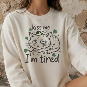 Op de afbeelding: Crèmekleurige sweatshirt met een illustratie van een chagrijnige kat met een hoed met een klavertje vier. De tekst "kiss me I'm tired" staat onder de kat, omringd door klaverafbeeldingen. Een kledingstuk met het thema St. Patrick's Day.