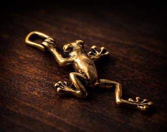 May include: A detailed, golden frog pendant with a loop for a chain. The frog is in a dynamic pose, with its legs extended and arms raised. The pendant is made of a shiny, metallic material and rests on a dark wooden surface.