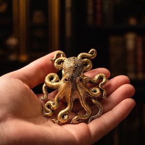 May include: A detailed, golden octopus figurine with textured skin and curled tentacles. The sculpture is held in an open hand, showcasing its intricate design and metallic sheen. The octopus is a decorative item.
