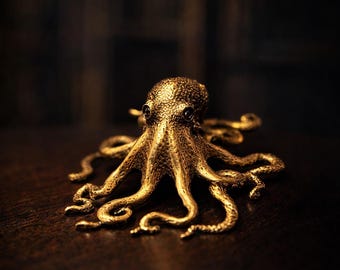 Può includere: Una scultura dettagliata di un polpo dorato, con pelle strutturata e occhi scuri. Gli otto tentacoli si arricciano con grazia e la scultura poggia su una superficie di legno scuro. L'estetica generale è elegante e dettagliata.
