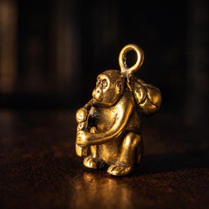 May include: A small, detailed brass monkey figurine, seated and holding an object, with a loop for hanging. The monkey has a bag over its shoulder. The figurine is golden in color and sits on a dark surface.