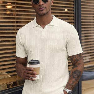 May include: A short-sleeved, cream-colored polo shirt featuring a vertical ribbed texture, a classic collar, and a buttoned placket. The shirt is worn with light-colored trousers, a silver-toned wristwatch, and dark-tinted round sunglasses.