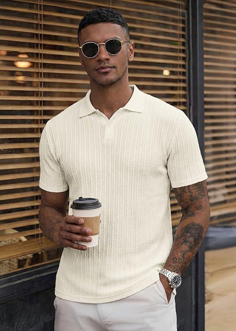 May include: A short-sleeved, cream-colored polo shirt featuring a vertical ribbed texture, a classic collar, and a buttoned placket. The shirt is worn with light-colored trousers, a silver-toned wristwatch, and dark-tinted round sunglasses.