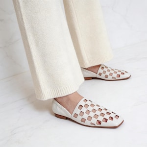 Può includere: Ballerine in pelle bianca con punta quadrata e design traforato. Le scarpe hanno un tacco basso e sono indossate con pantaloni color crema. Le ballerine sono un'opzione di calzatura elegante e confortevole.