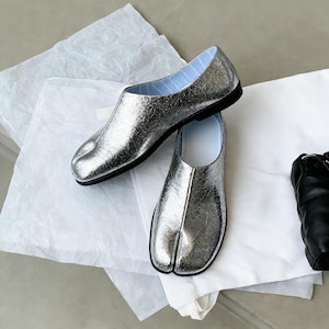 Puede incluir: Un par de zapatos plateados con puntera dividida y suelas negras, sobre papel blanco. Los zapatos tienen un aspecto met&aacute;lico texturizado y un forro a rayas azul claro. Un dise&ntilde;o &uacute;nico.