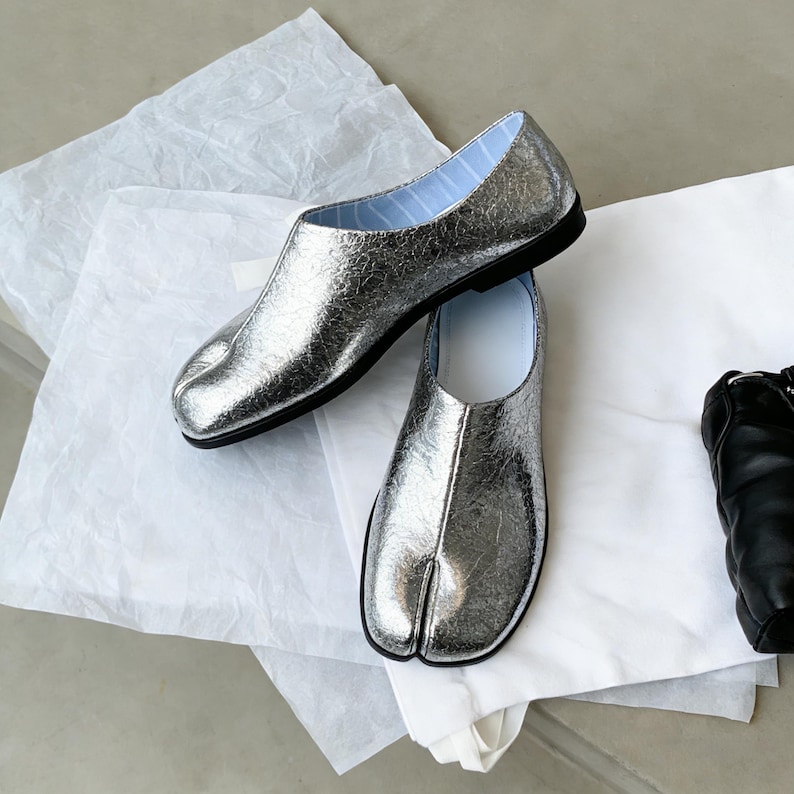 Puede incluir: Un par de zapatos plateados con puntera dividida y suelas negras, sobre papel blanco. Los zapatos tienen un aspecto met&aacute;lico texturizado y un forro a rayas azul claro. Un dise&ntilde;o &uacute;nico.