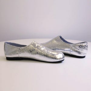 Puede incluir: Un par de zapatos plateados sin cordones con un acabado met&aacute;lico texturizado. Los zapatos tienen una puntera redondeada y una suela baja negra. El forro interior presenta un estampado de rayas azules y blancas.