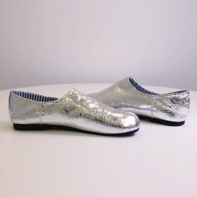Puede incluir: Un par de zapatos plateados sin cordones con un acabado met&aacute;lico texturizado. Los zapatos tienen una puntera redondeada y una suela baja negra. El forro interior presenta un estampado de rayas azules y blancas.