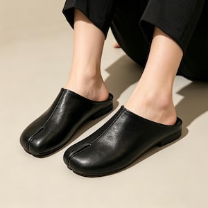 May include: Black leather slip-on shoes with a split-toe design. The shoes have a low heel and a smooth, polished finish. The footwear is designed for comfort and style.