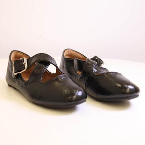 Peut inclure: Paire de ballerines noires de style Mary Jane avec une finition texturée. Chaque chaussure est dotée d'une bride sur le dessus avec une boucle argentée. Les chaussures ont un bout arrondi et un petit talon.