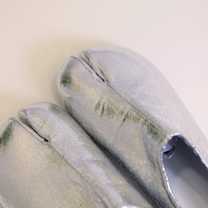 Puede incluir: Un par de zapatos plateados con punta dividida y apariencia agrietada y texturizada. Los zapatos tienen un dise&ntilde;o &uacute;nico, con una separaci&oacute;n para el dedo gordo. El acabado met&aacute;lico les da un aspecto moderno y llamativo.
