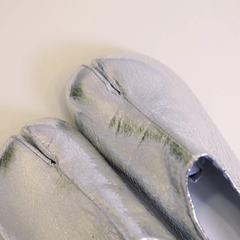 Puede incluir: Un par de zapatos plateados con punta dividida y apariencia agrietada y texturizada. Los zapatos tienen un dise&ntilde;o &uacute;nico, con una separaci&oacute;n para el dedo gordo. El acabado met&aacute;lico les da un aspecto moderno y llamativo.