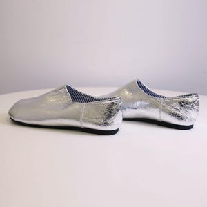 Puede incluir: Un par de zapatos plateados sin cordones con un acabado texturizado. Los zapatos tienen suela negra y forro a rayas azules y blancas visible en la abertura. Los zapatos se exhiben sobre una superficie blanca.