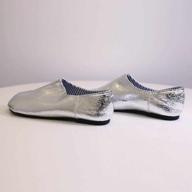 Puede incluir: Un par de zapatos plateados sin cordones con un acabado texturizado. Los zapatos tienen suela negra y forro a rayas azules y blancas visible en la abertura. Los zapatos se exhiben sobre una superficie blanca.