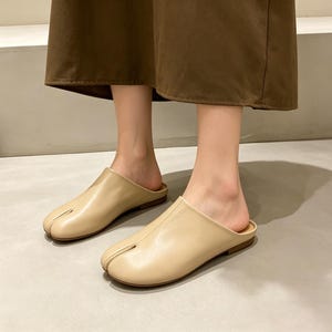 May include: Beige leather slip-on shoes with a split toe design. The shoes have a low heel and a minimalist aesthetic. The footwear is shown from a low angle, highlighting the shape and style of the shoes.