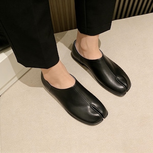 May include: A pair of black leather split-toe shoes. The shoes have a unique design with a separate compartment for the big toe. The shoes are worn by a person wearing black pants.